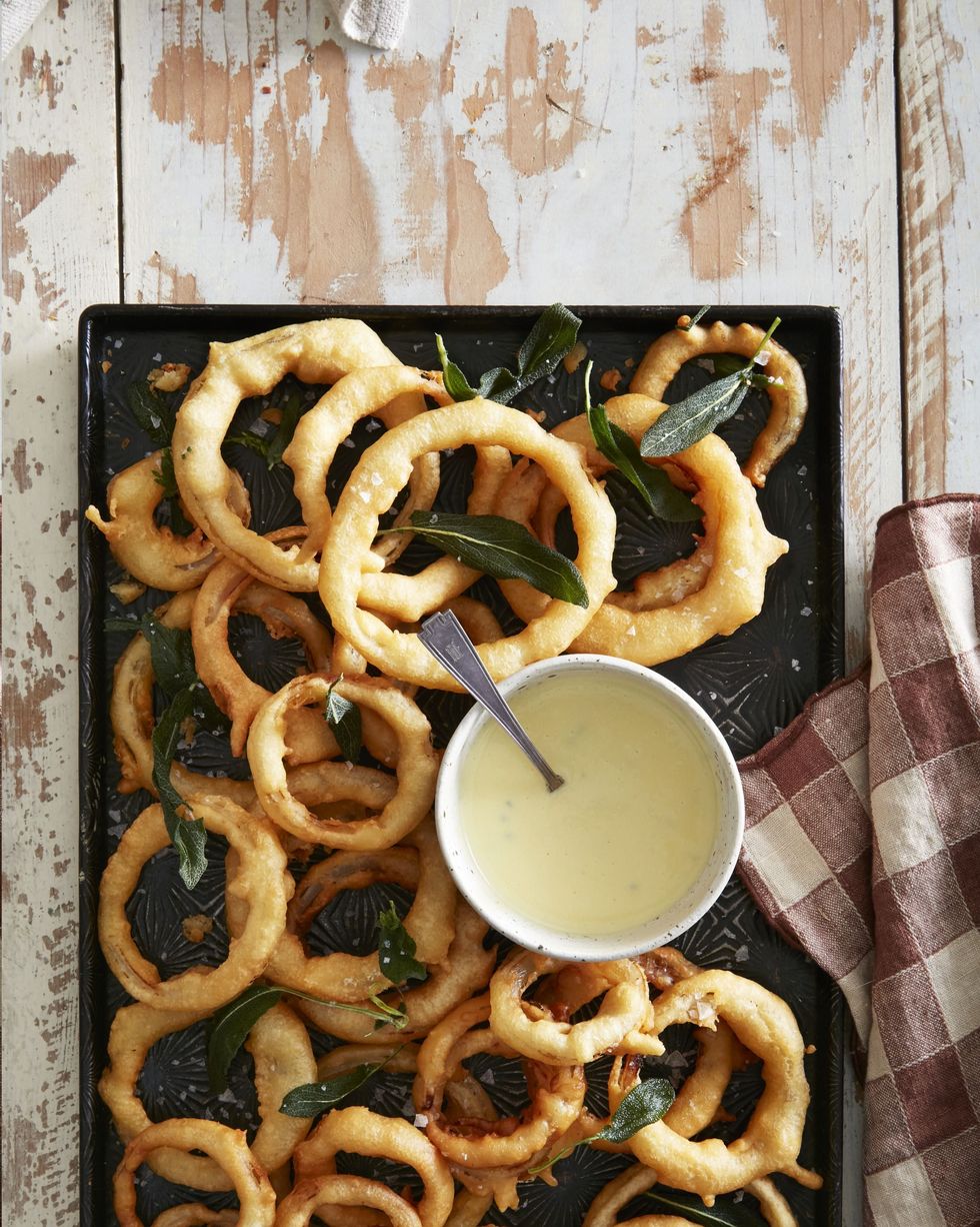 onion rings on a sheet pan with a bowl of sage aioli and a spoon for serving