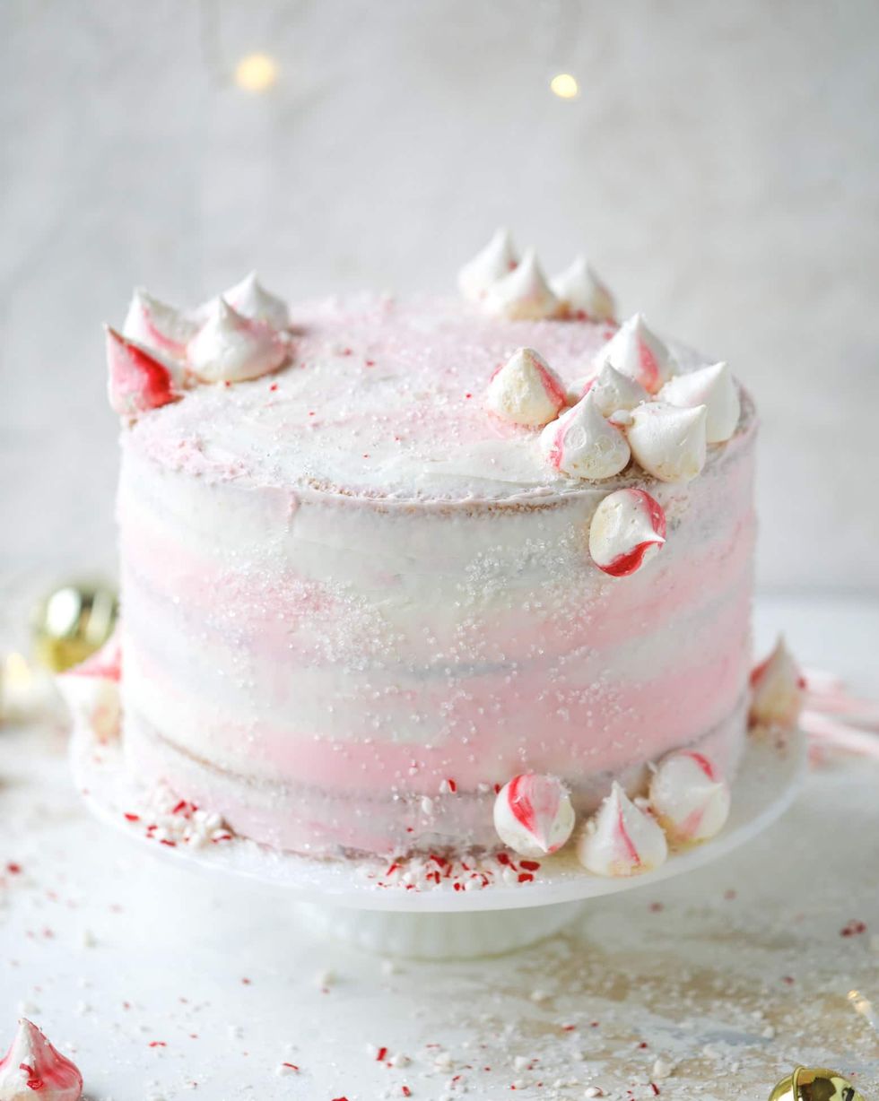 layer cake covered in white and light pink swirled frosting and garnished with sugar sprinkles and red and white swirled meringue kisses