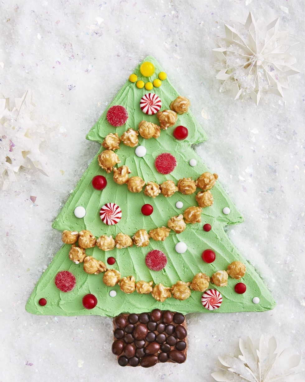 a sheet cake cut into the shape of a christmas tree with brown frosting for the stump and green frosting for the leaves and caramel corn for garland and carious white and red candies for ornaments
