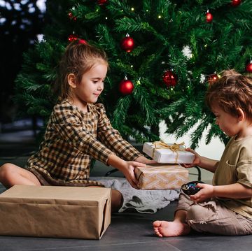 diligent little siblings sitting on the floor under a decorated christmas tree, holding wrapped presents