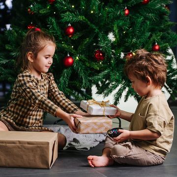 diligent little siblings sitting on the floor under a decorated christmas tree, holding wrapped presents