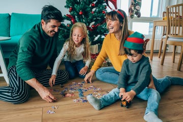 two cute children and their mother and father sitting on the floor near the christmas tree and arranging a jigsaw puzzle