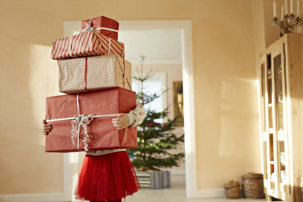 little girl holding tall stack of christmas presents, standing in living room