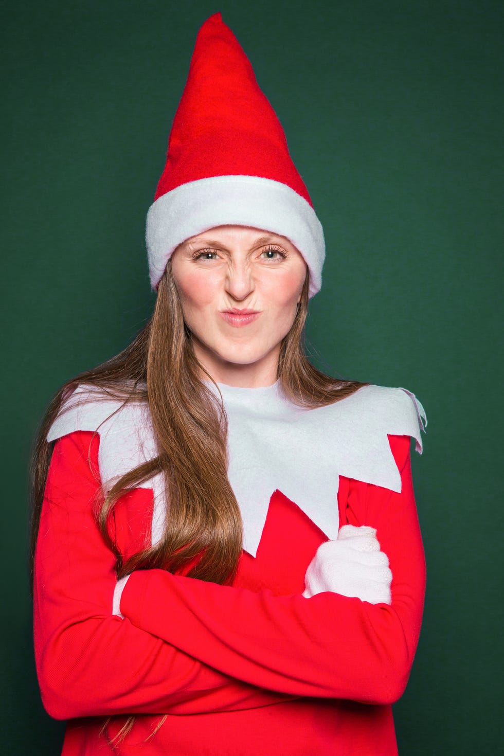 young woman wearing elf costume and red hat standing with arms crossed against a dark green background