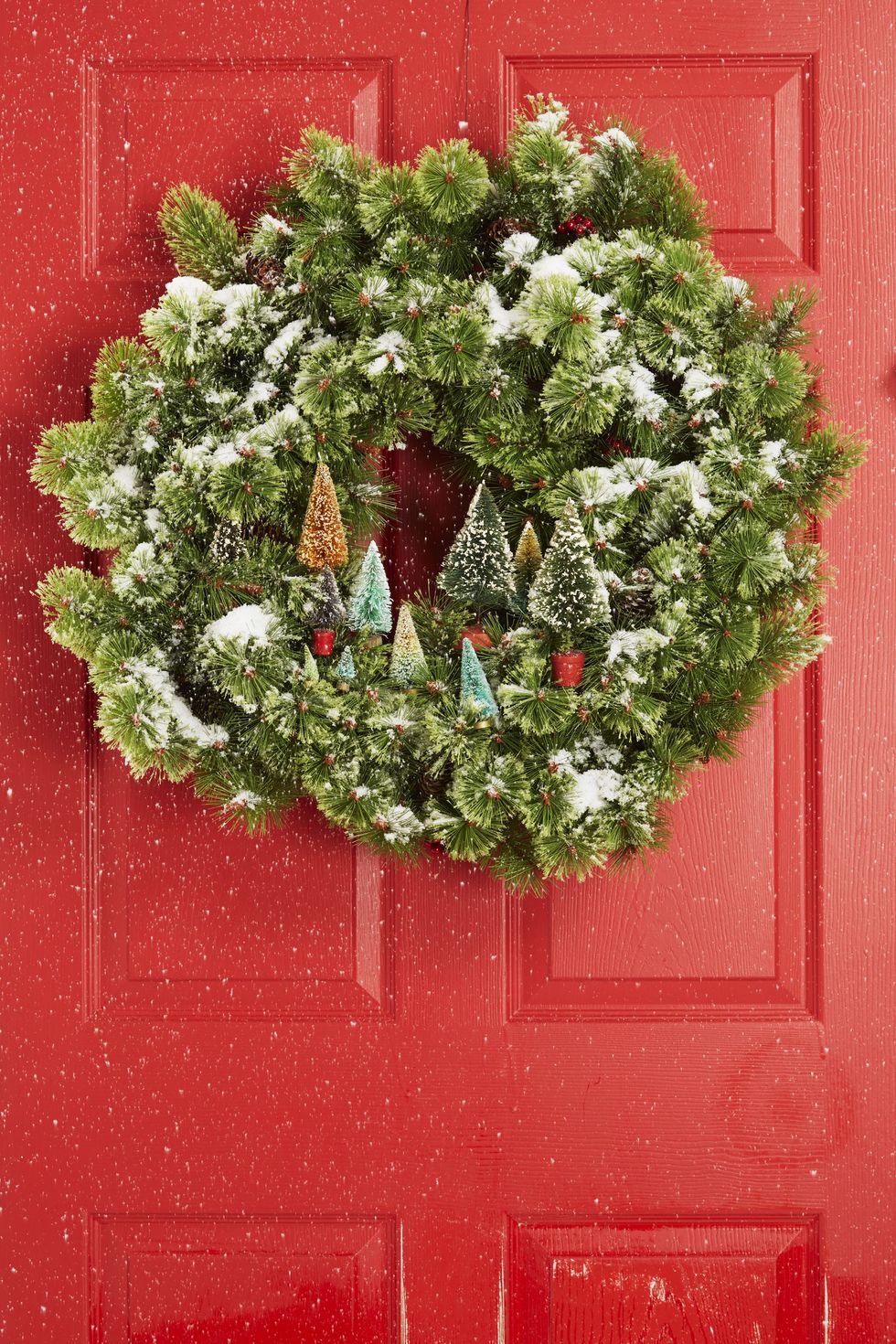 diy christmas wreath, greenery wreath with fake snow and small fake christmas trees hanging from a red door
