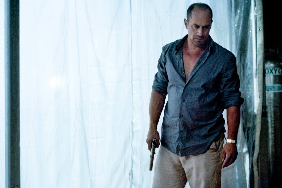 christopher meloni holding a firearm wearing a partially unbuttoned shirt and beige trousers set against a backdrop of a white curtain in carriers