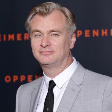 christopher nolan looks at the camera while standing in front of a dark blue background, he wears a gray suit jacket, white collared shirt and black tie