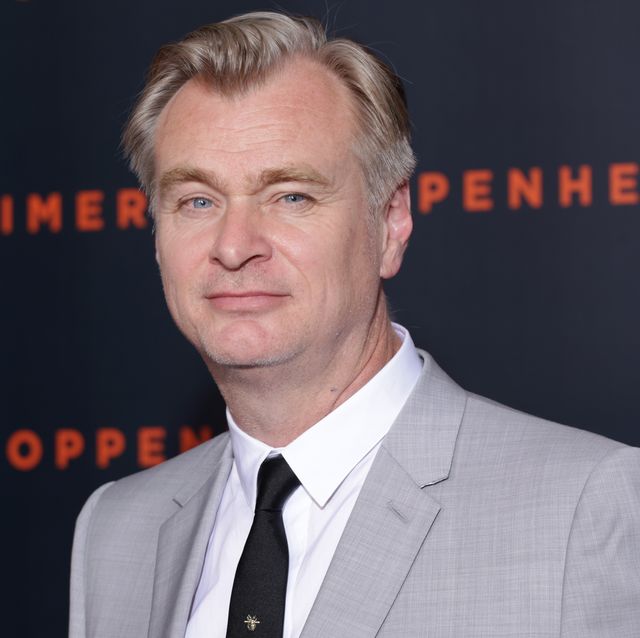 christopher nolan looks at the camera while standing in front of a dark blue background, he wears a gray suit jacket, white collared shirt and black tie