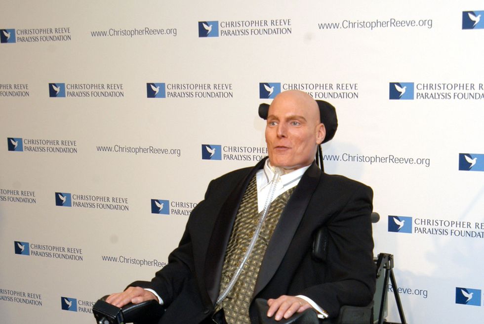 a man in a wheelchair sits in a three piece suit in front of a white background with a logo