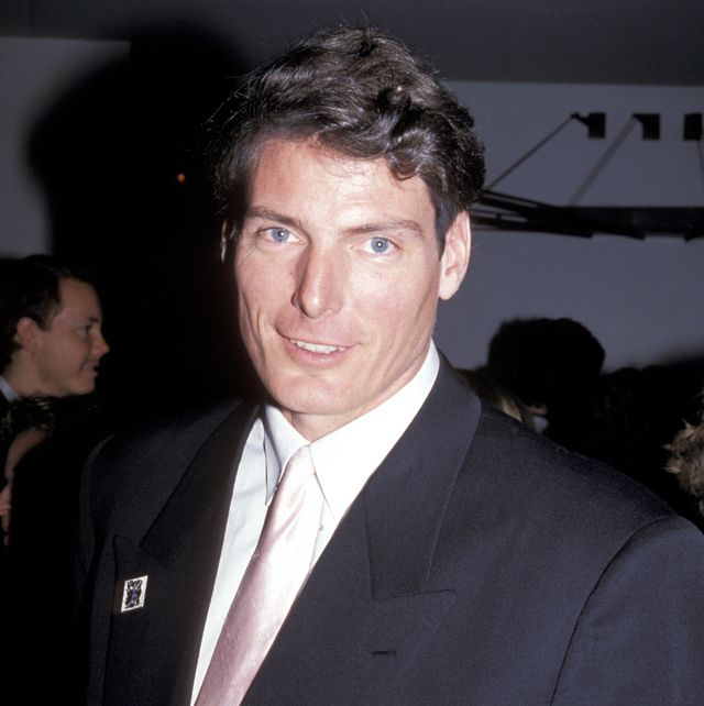 christopher reeve smiles at the camera, he wears a black suit jacket with a white collared shirt and light pink tie