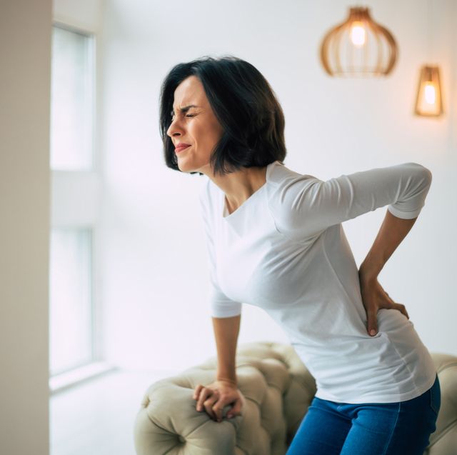 chronic back pain adult woman is holding her lower back, while standing and suffering from unbearable pain