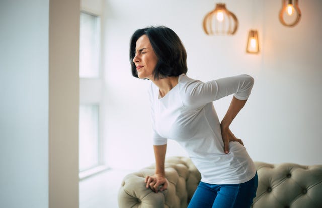 chronic back pain adult woman is holding her lower back, while standing and suffering from unbearable pain