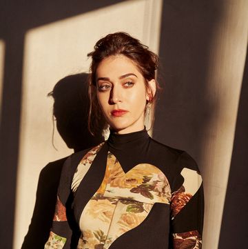 actor lizzy caplan shown in a dramatic portrait with her hair up, eyebrow arched, in a black and floral bodysuit tucked into jeans