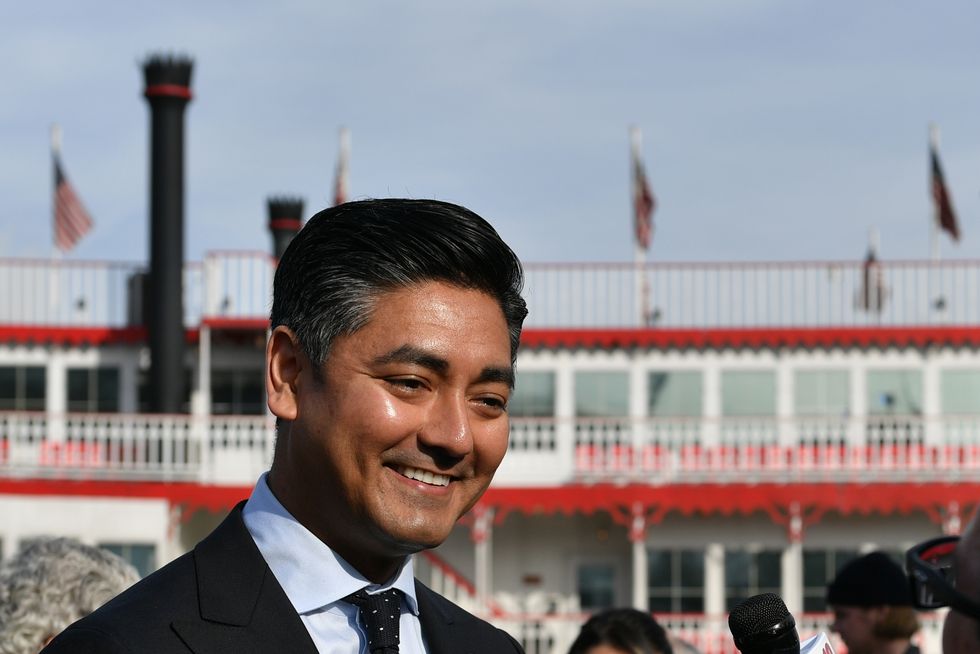 cincinnati major aftab pureval smiles as unseen people hold microphones toward him, he wears a suit and tie and stands outside