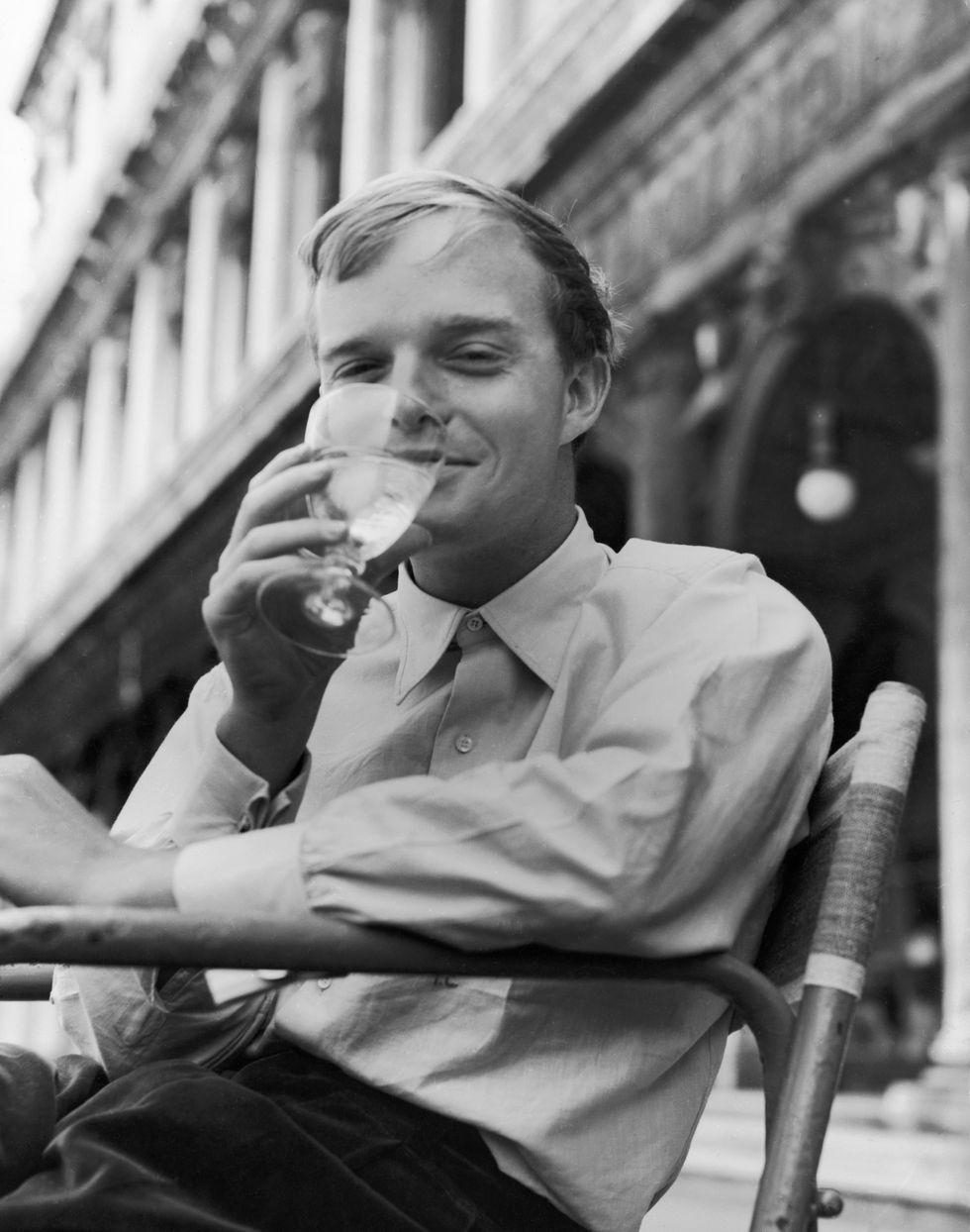truman capote smiles while taking a drink from a glass, he sits outside a stone building and wears a collared shirt and pants