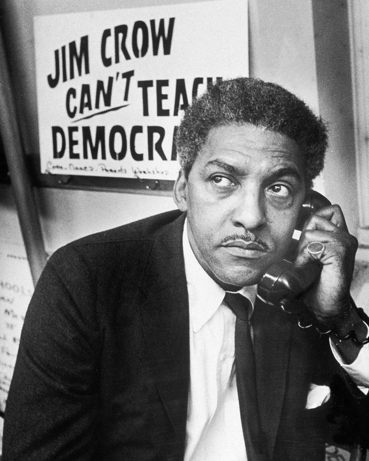 bayard rustin holds a telephone to one ear and looks to the left, behind him is a protest sign, he wears a suit and tie