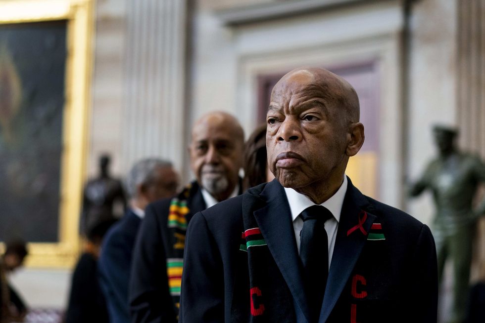 john lewis stands inside a large room in a blue suit jacket and tie, he also wears a black scarf with red letters and a red and green stripe, he looks to the left and people stand behind