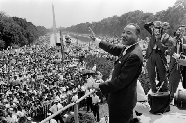 martin luther king jr, waving and smiling, stands in a suit on a platform, crowds of people look on from the background, the washington monument and reflection pool are in the background too, two cameramen stand on the right