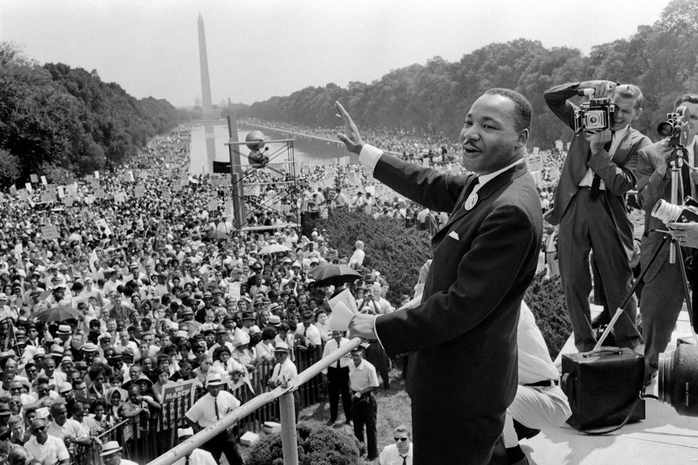 martin luther king jr, waving and smiling, stands in a suit on a platform, crowds of people look on from the background, the washington monument and reflection pool are in the background too, two cameramen stand on the right