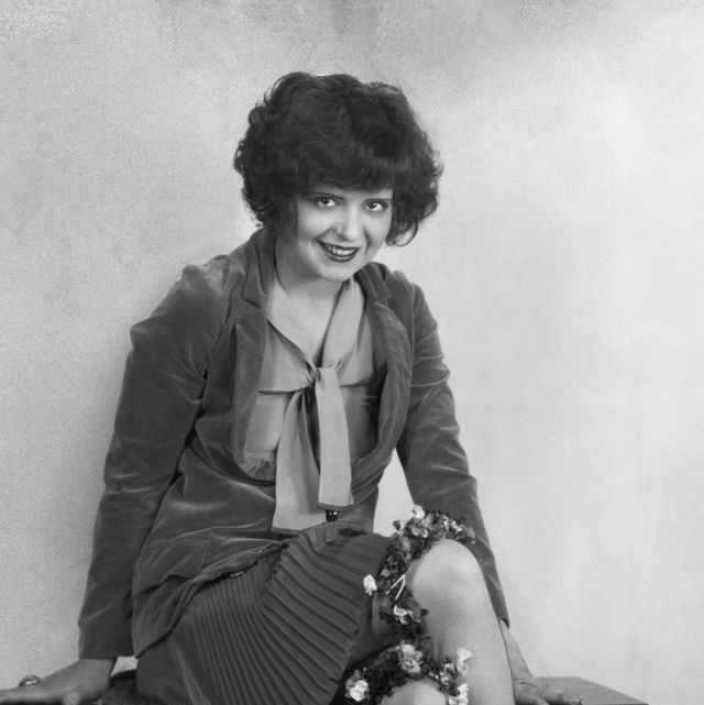clara bow sits on a bench and smiles at the camera, she wears a jacket over a blouse with a pleated skirt and flower garters