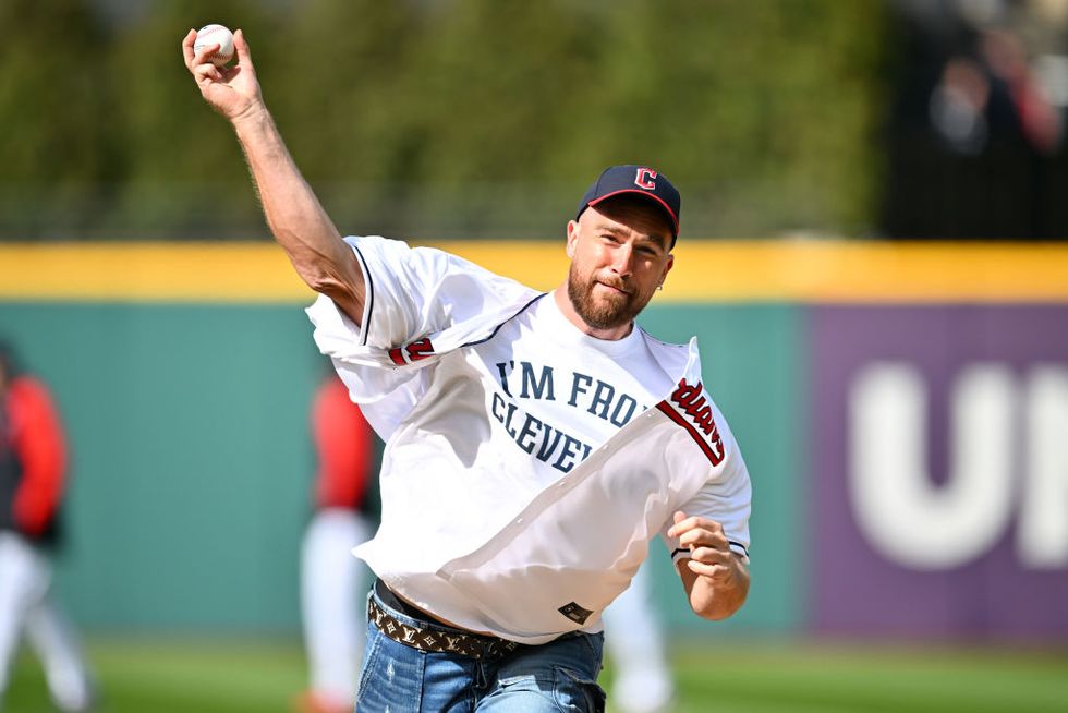 travis kelce, wearing a shirt that says im from cleveland underneath a cleveland guardians jersey, throws a baseball at a baseball field