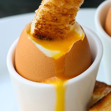 close up image of white buttered toasted bread slice being dipped into runny yellow yolk of soft boiled egg, toast soldiers with two soft boiled eggs on white plate background, focus on foreground