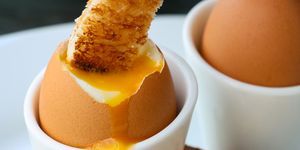 close up image of white buttered toasted bread slice being dipped into runny yellow yolk of soft boiled egg, toast soldiers with two soft boiled eggs on white plate background, focus on foreground