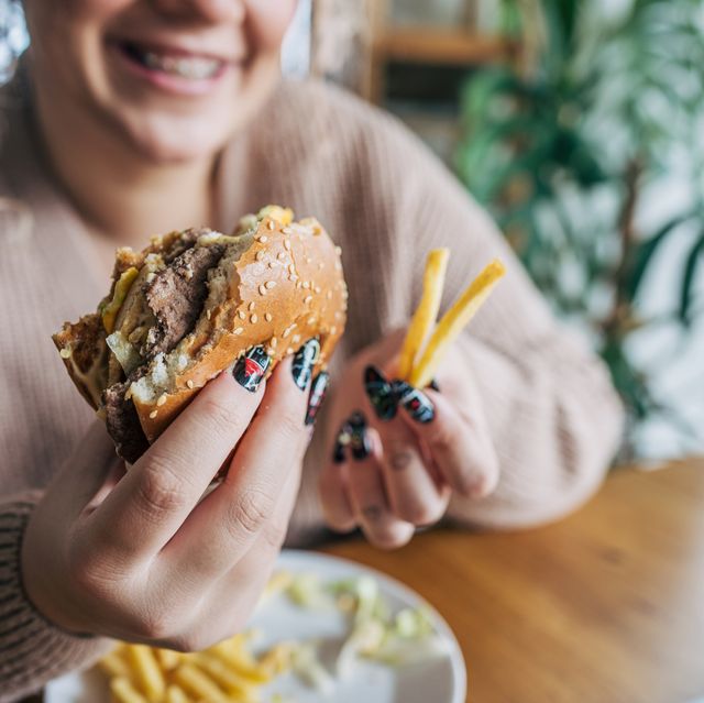close up of a woman with curly hair sitting at a wooden dining table in a cozy, well lit room, holding a burger and fries in her hand