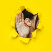 close up of a woman's ear and hand through a torn hole in the paper bright yellow background, copy space the concept of eavesdropping, espionage, gossip and tabloids