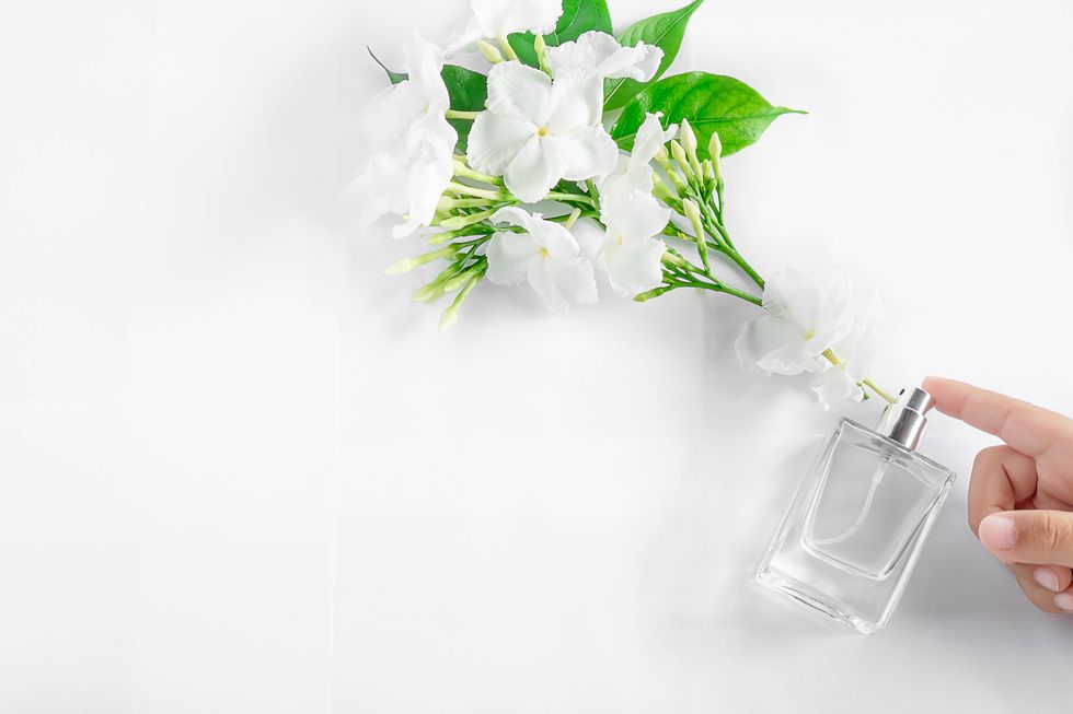 Close-Up Of Hand Touching Perfume Bottle By Flowers Over White Background