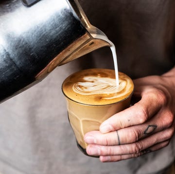 Close up of person with tattooed finger pouring milk from metal jug into glass of cafe latte.