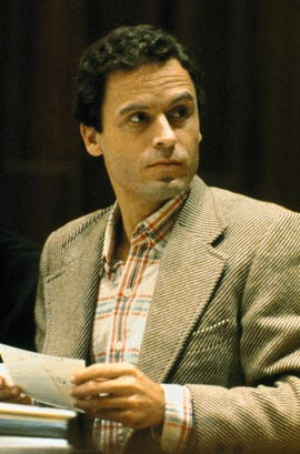 ted bundy looks over his shoulder with a neutral expression on his face, he wears a tan and cream striped suit jacket with a red, white, and blue collared shirt