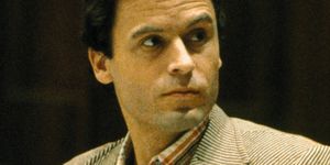 ted bundy looks over his shoulder with a neutral expression on his face, he wears a tan and cream striped suit jacket with a red, white, and blue collared shirt