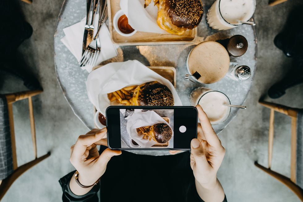 close up of woman's hand taking photo of freshly made burger with french fries by smartphone while having meal with friends in restaurant