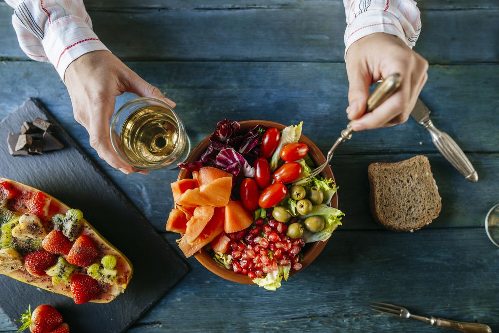 Close-up of woman's hands eating salad wit tomato, pomegranate, papaya and olives, papaya with fruits and with wine glass