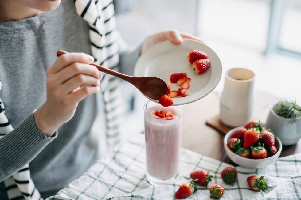 close up of young asian woman making fresh and healthy strawberry smoothie for breakfast, adding chopped strawberries on top as garnish healthy eating and healthy lifestyle