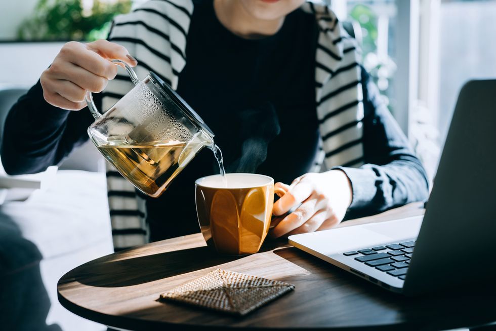 close up of young asian woman pouring a cup of tea from a stylish transparent tea pot into a cup starting a great day ahead with a cup of hot tea while working at home on laptop in the fresh morning against sunlight healthy lifestyle concept