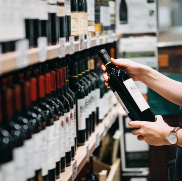 close up of young asian woman walking through supermarket aisle and choosing a bottle of red wine from the shelf in a supermarket