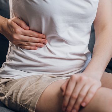 close up of young female with fatty liver touches right side with hand, suffering from abdominal pain sit on grey couch pain in right side, appendix, gallstones and gynecological diseases concept