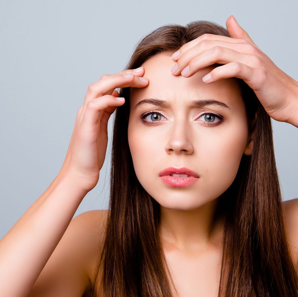 Close up portrait of frustrated sad upset beautiful young woman is squeezing out pimples on her forehead, isolated on grey background