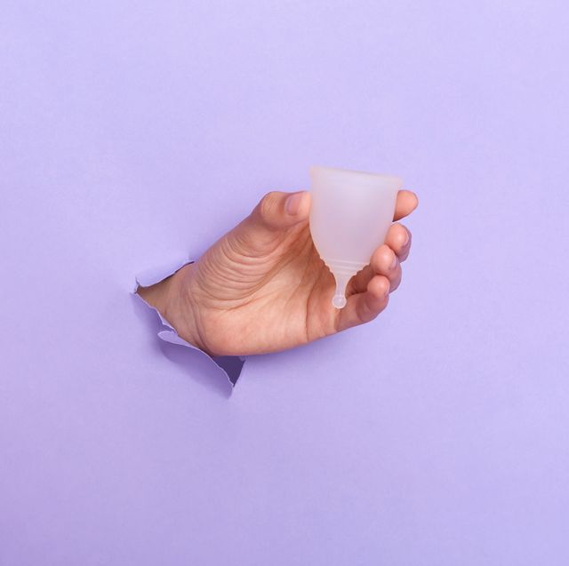 close up shot of human's hand holding menstrual cup in hole of purple paper wall, modern sanitary products for period, women health, gynecology, hygiene, zero waste alternatives