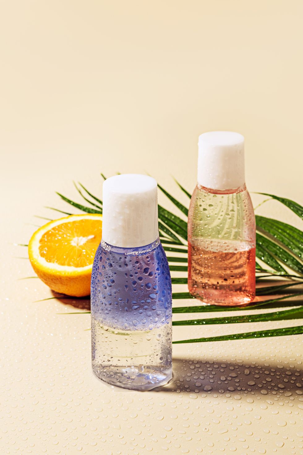close up view of micellar water in bottles, green palm leaf and orange half with water drops on beige backdrop