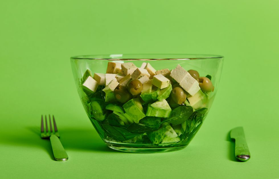 closeup of a serving bowl with green salad and vegetables, and vegan tofu cheese on green background