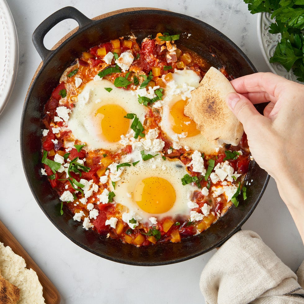 closeup of female hand dunking flatbread in shakshuka in frying pan