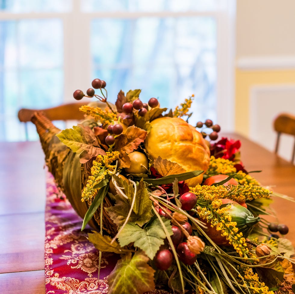 a closeup of thanksgiving fake cornucopia decoration filled with leaves and fruit and bread on a wooden table with a red tablecloth