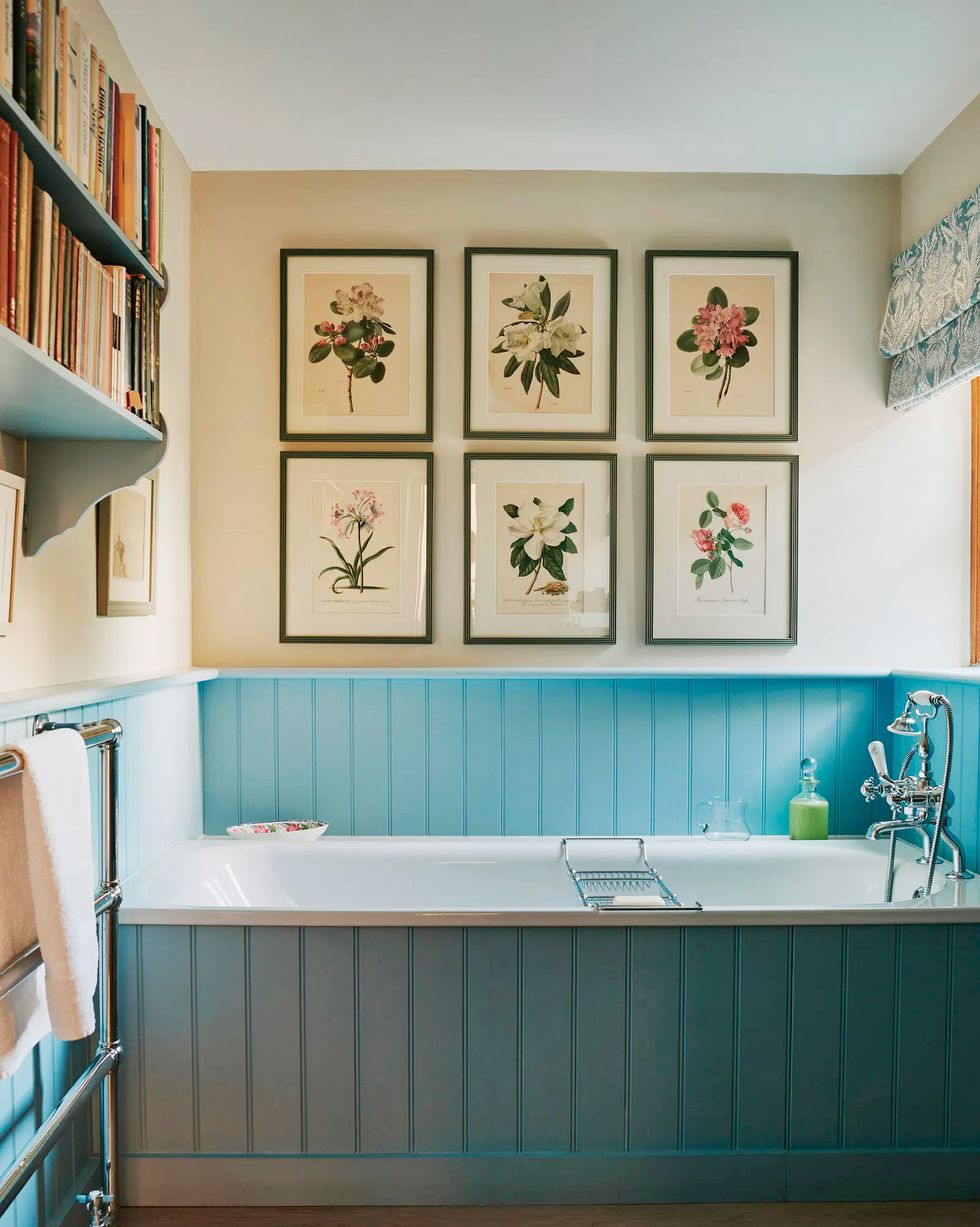 botanical prints hang above a bathtub with a blue wood surround in a country home