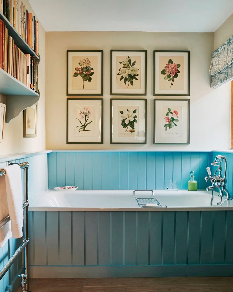 botanical prints hang above a bathtub with a blue wood surround in a country home