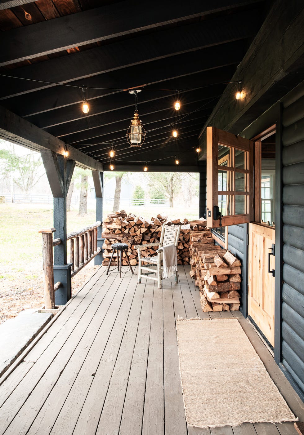 front porch of a 1970s black log cabin in upstate new york features a white wood chair and piles of firewood