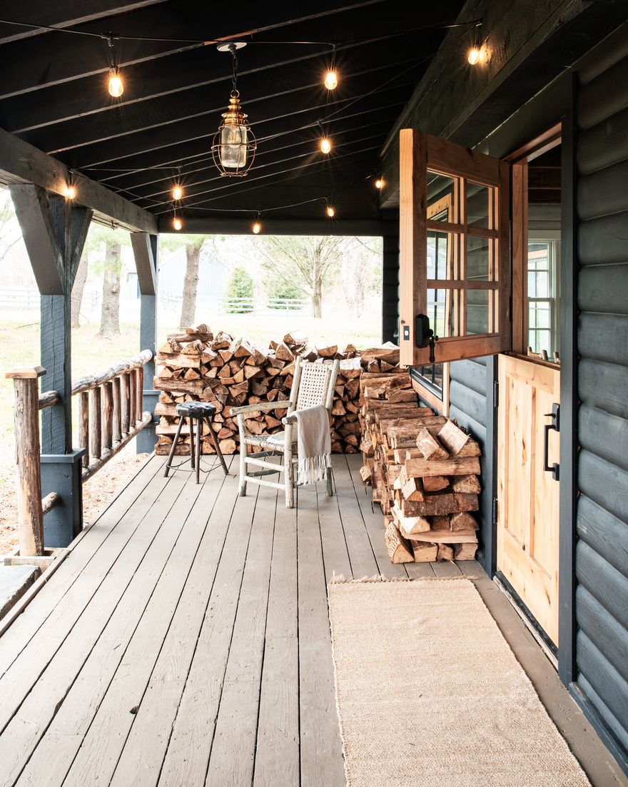 front porch of a 1970s black log cabin in upstate new york features a white wood chair and piles of firewood