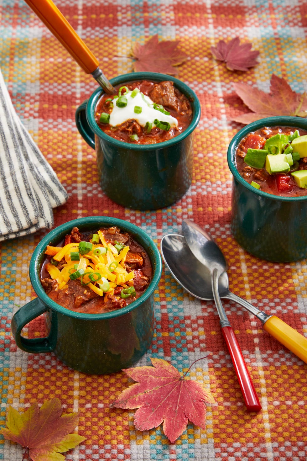 beef chili with fixins in green enamelware mugs set on a plaid tablecloth